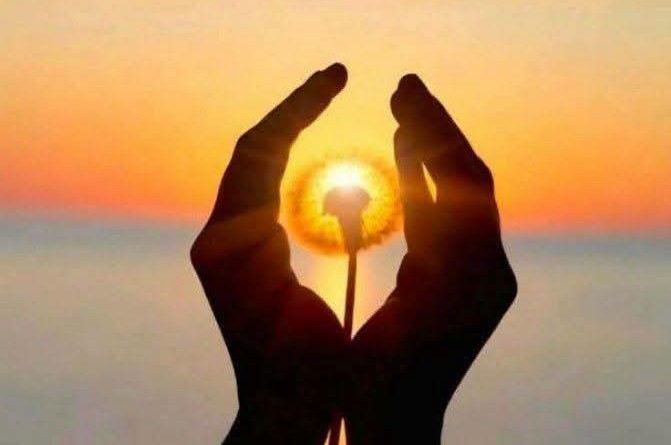 Twee handen vormen een hartvorm rond een brandende fakkel tegen een oranje zonsondergang.
